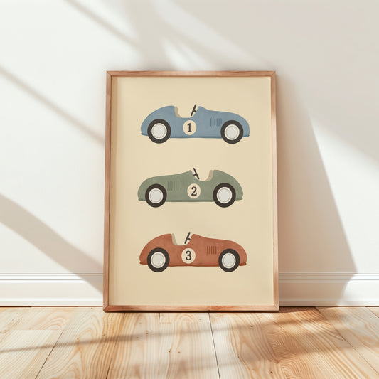 A framed art print featuring three retro race cars in blue, green, and brown on a cream background, leaning against a white wall on a light wood floor.