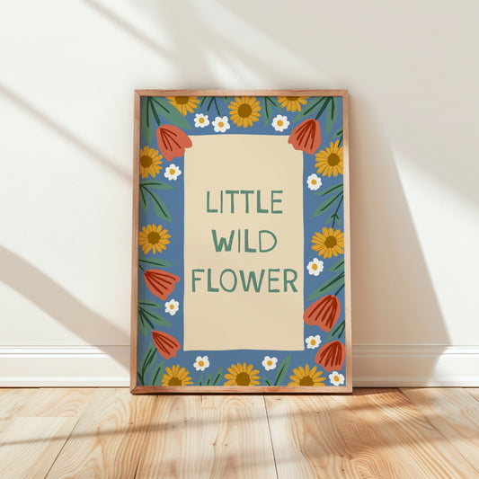 A framed art print with the text 'LITTLE WILD FLOWER' in green on a cream background, surrounded by a blue border with yellow sunflowers, white daisies, and orange-red tulips. The frame is leaning against a white wall on a light wood floor with soft diagonal shadows.