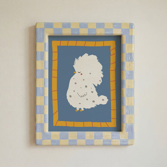 A framed art print featuring a cute white fluffy bird with small gray spots on a dusty blue background, surrounded by a yellow-orange patterned border and a blue-and-beige checkered frame, mounted on a light beige wall.