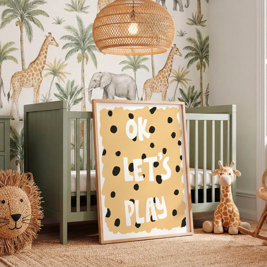 A styled nursery room featuring a sage green wooden crib, a large framed 'OK LET'S PLAY' sign with black polka dots on a yellow background, safari-themed wallpaper with giraffes and palm trees, a woven rattan pendant light, and a plush giraffe toy.