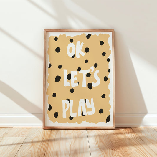 A framed typographic art print leaning against a white wall on a light wood floor. The print features the words 'OK LET'S PLAY' in white hand-drawn letters on a warm beige background with black polka dots, enclosed in a light natural wood frame.