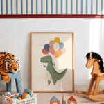 A framed children's art print featuring a green dinosaur holding colorful balloons, styled in a nursery or playroom setting with a tiger plush, wooden rocking horse, and toys on the floor.