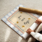 A person unrolling a decorative wall mural or poster on a beige carpet. The design features a central cream panel with the words 'you are Magic' in black script, surrounded by gold stars and scalloped pastel pink and blue borders, with a blue-and-white diamond pattern around the outer edge.