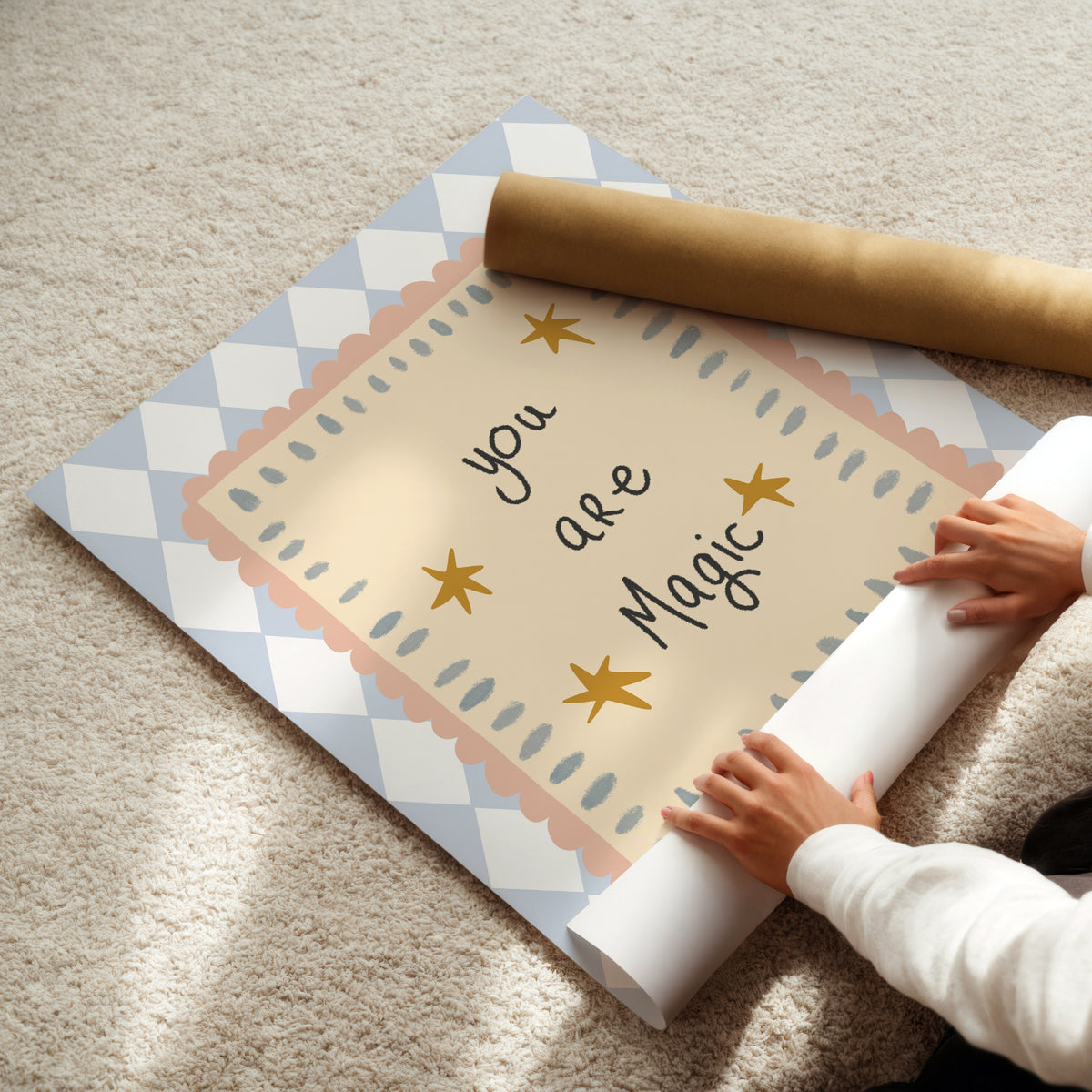 A person unrolling a decorative wall mural or poster on a beige carpet. The design features a central cream panel with the words 'you are Magic' in black script, surrounded by gold stars and scalloped pastel pink and blue borders, with a blue-and-white diamond pattern around the outer edge.