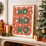 Framed Christmas art print leaning on a fireplace mantel, featuring the words 'HO HO HO' in green on a terracotta background with white polka dots. The scene is decorated with wrapped gifts, pinecones, a Christmas tree, and a white brick fireplace.