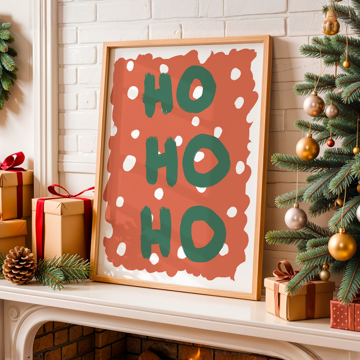 Framed Christmas art print leaning on a fireplace mantel, featuring the words 'HO HO HO' in green on a terracotta background with white polka dots. The scene is decorated with wrapped gifts, pinecones, a Christmas tree, and a white brick fireplace.