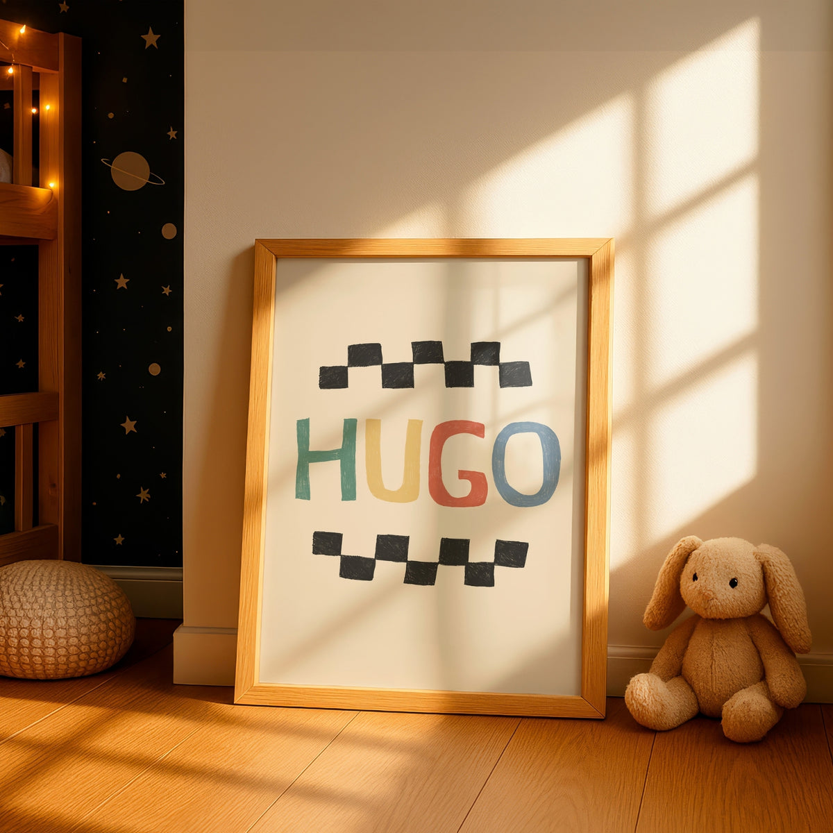 A framed children's wall art print leaning against a wall in a sunlit nursery. The print has the name 'HUGO' in multicolored letters between black checkered lines, with a light wood frame.