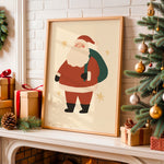 A framed Christmas art print featuring a cartoon Santa Claus with a green sack, leaning on a fireplace mantel next to a decorated Christmas tree and wrapped gift boxes.