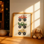 A framed children's art print leaning against a wall in a styled nursery. The print features three monster trucks in red, green, and tan on an off-white background, enclosed in a light wood frame.
