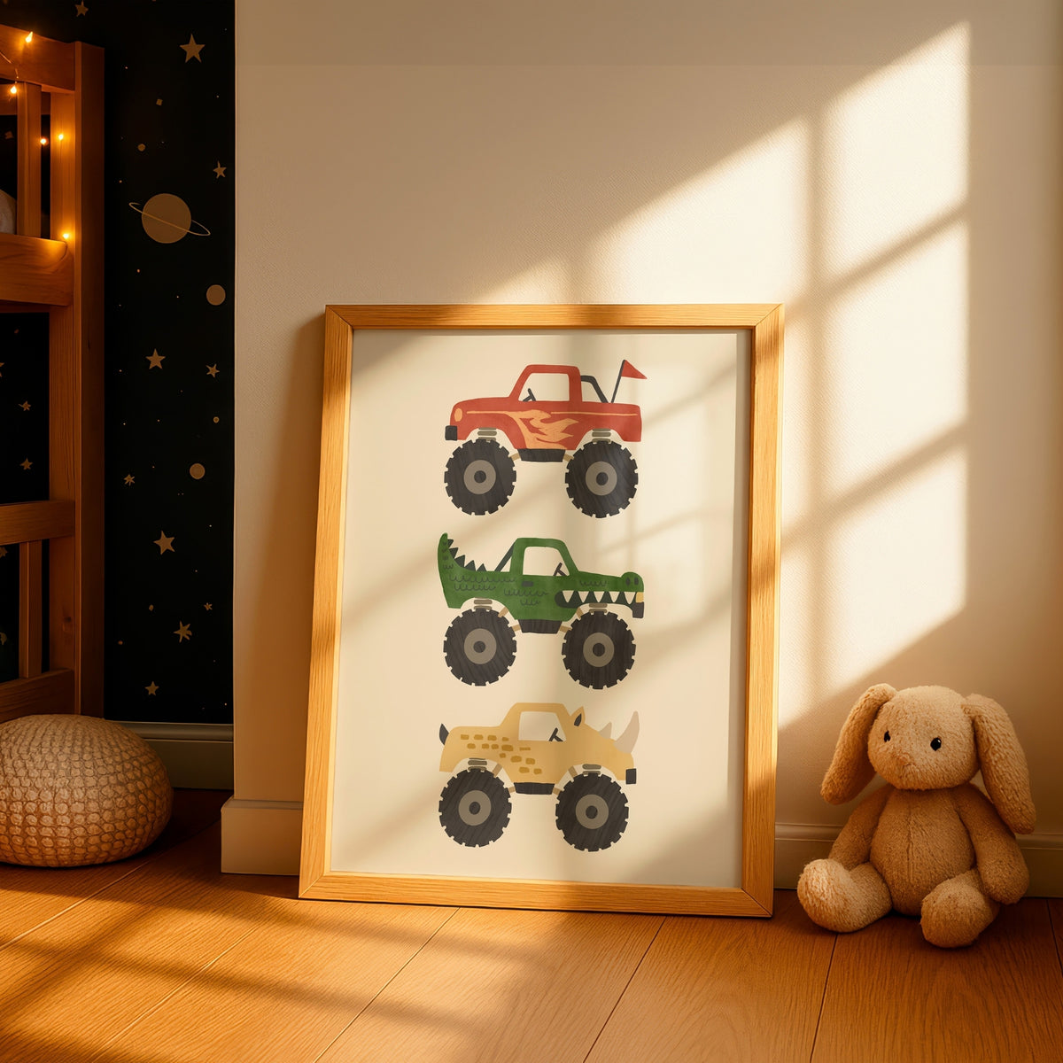 A framed children's art print leaning against a wall in a styled nursery. The print features three monster trucks in red, green, and tan on an off-white background, enclosed in a light wood frame.