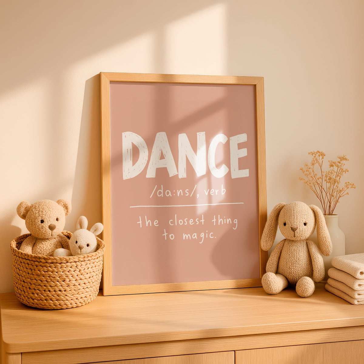 A framed nursery-style wall print with the word "DANCE" in large white letters on a dusty pink background, styled on a wooden dresser beside plush toys and dried flowers.