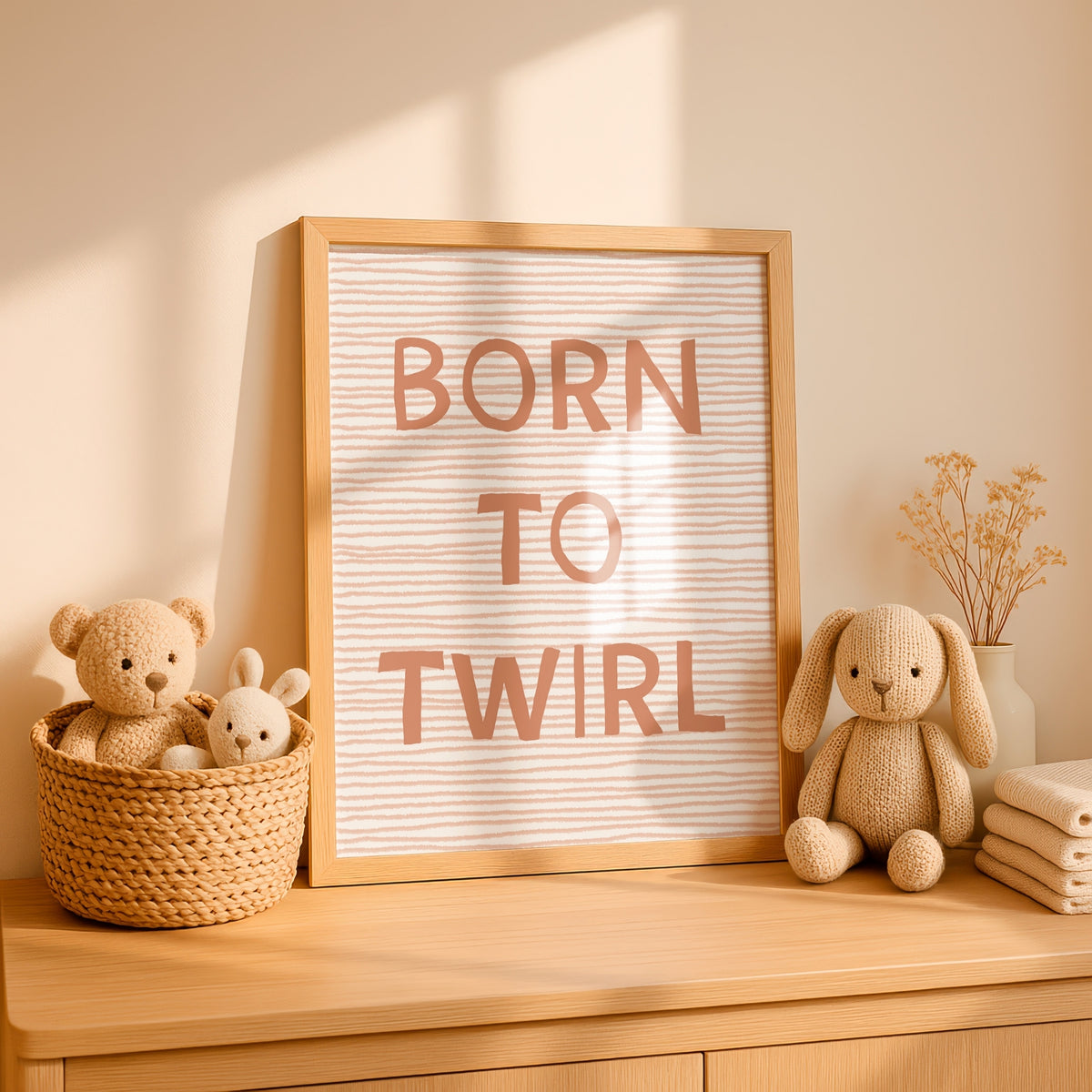 A framed nursery wall sign with the words “BORN TO TWIRL” in dusty pink lettering on a white striped background, styled on a wooden dresser with plush toys and dried flowers in a warm, sunlit room.