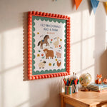 A framed children's educational poster reading 'OLD MACDONALD HAD A FARM' hangs on a wall above a wooden table with pencils, a globe, and small toys. The poster has a coral pom-pom border, a teal inner border, and illustrated farm animals on a white background.