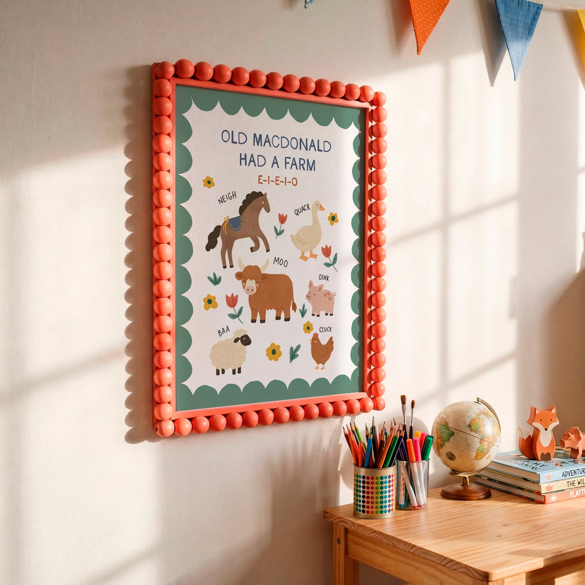 A framed children's educational poster reading 'OLD MACDONALD HAD A FARM' hangs on a wall above a wooden table with pencils, a globe, and small toys. The poster has a coral pom-pom border, a teal inner border, and illustrated farm animals on a white background.