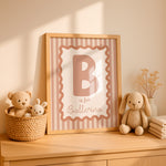 A framed nursery wall art print leaning on a wooden dresser, featuring a large letter B and the words 'is for Ballerina' on a pink striped background. The scene includes a woven basket with teddy bears, a knitted bunny, dried flowers, and folded blankets in a warm, sunlit room.