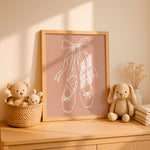 A framed art print featuring white line art of ballet pointe shoes on a dusty pink background, styled on a light wood dresser next to plush toys and dried flowers.