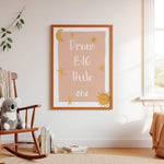 A framed nursery wall art print with the words 'Dream BIG little one' in white script on a dusty pink background, decorated with gold stars, a crescent moon, and a sun. The print is displayed in a light wood frame on a white wall in a styled nursery setting with a wooden rocking chair, a plush koala, and a window.