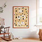 A framed wall art print with the words “OK LET’S PLAY” in white lettering on a warm beige background with black spots, styled in a bright nursery or children’s room above a wooden rocking chair with a plush toy.