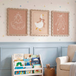 Three framed nursery wall art prints hang above a white book rack and beige armchair in a styled nursery room. The prints show simple line drawings of a ballerina, a duck, and ballet slippers on dusty pink backgrounds, with star decals on the wall.