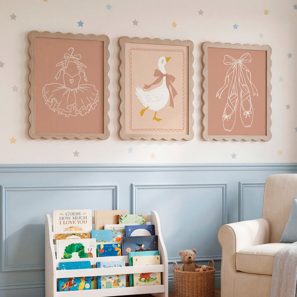 Three framed nursery wall art prints hang above a white book rack and beige armchair in a styled nursery room. The prints show simple line drawings of a ballerina, a duck, and ballet slippers on dusty pink backgrounds, with star decals on the wall.