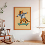 A framed art print featuring a cartoon tiger wearing a navy and white striped shirt and riding a green skateboard, displayed on a white wall in a styled nursery room with a wooden rocking chair, a plush toy, and books.