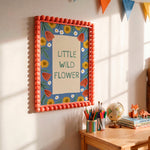A framed wall art print with the words “LITTLE WILD FLOWER” hangs on a sunlit cream wall above a wooden desk. The artwork has a coral-orange beaded border, a blue background with yellow sunflowers, white flowers, and orange leaves, plus a beige center panel with green lettering.