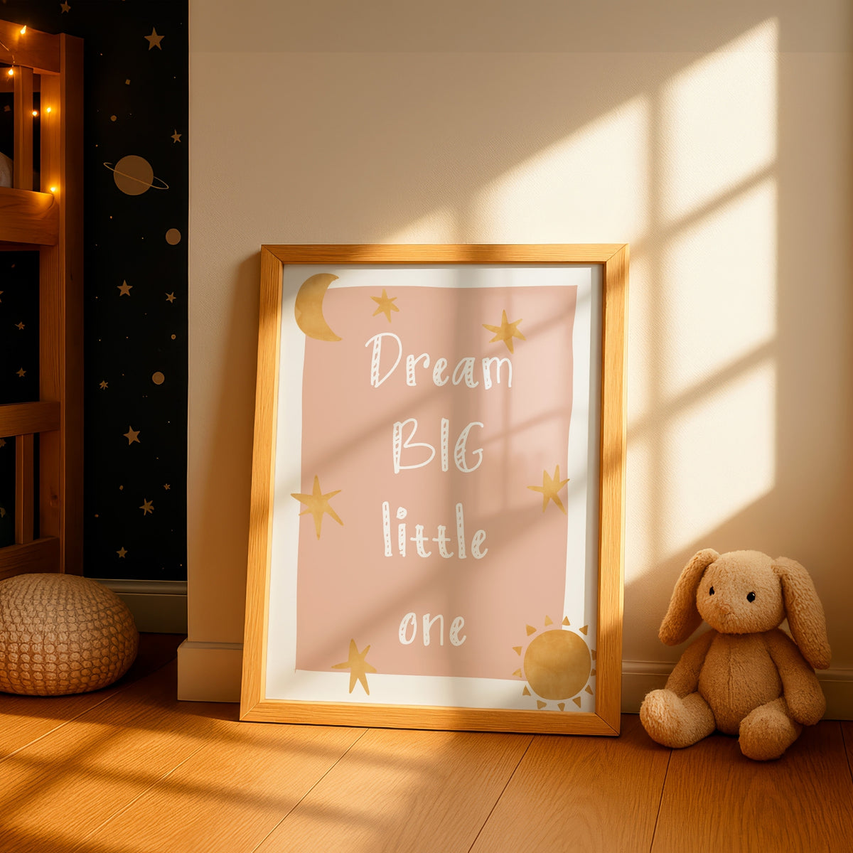 A framed nursery art print leaning against a wall in a sunlit room. The print has a pink background with white text reading 'Dream BIG little one' and gold stars, crescent moon, and sun illustrations.