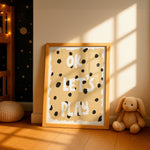 A framed children's wall art print leaning on a wooden floor in a sunlit nursery. The print has a warm beige background with black polka dots and the words 'OK LET'S PLAY' in white.