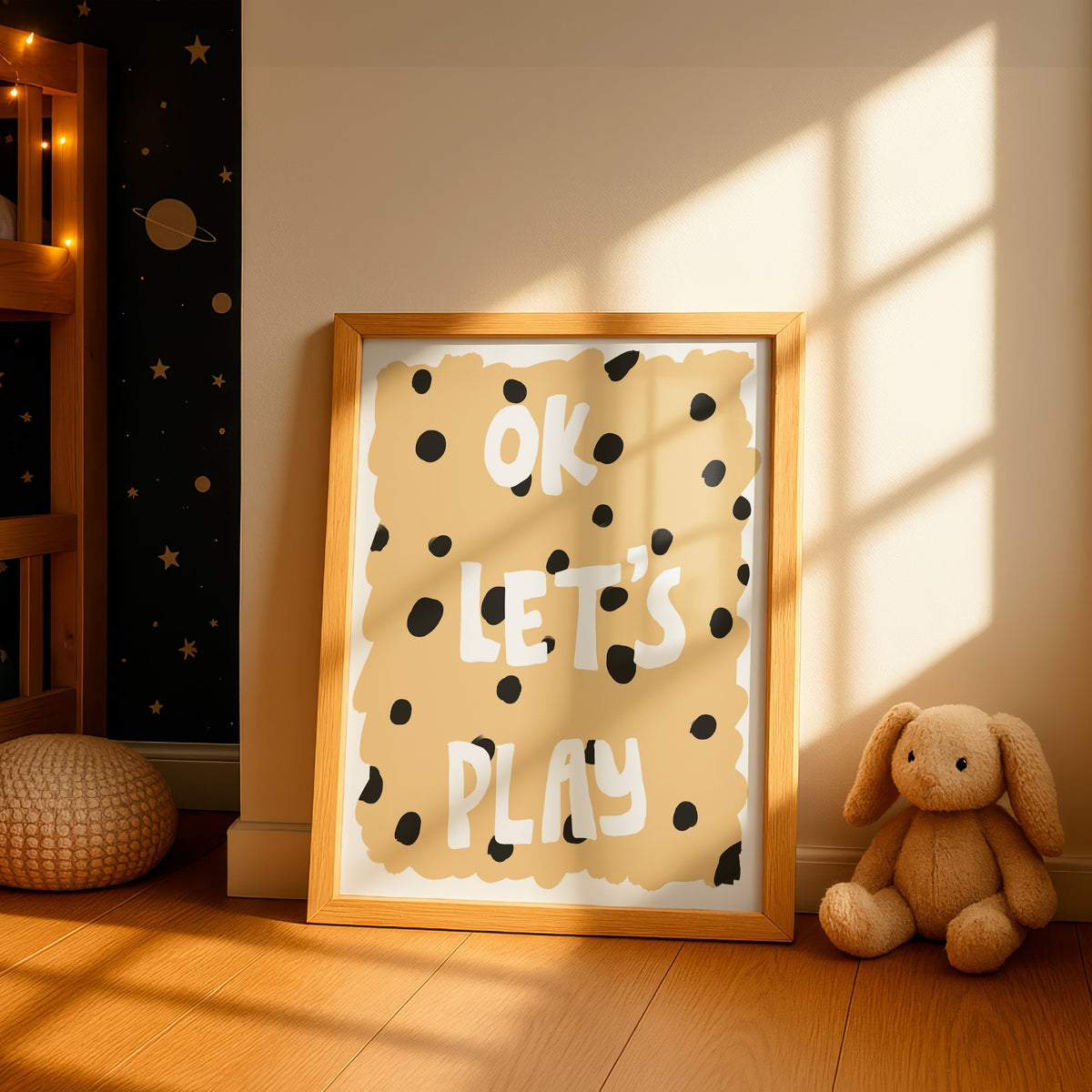 A framed children's wall art print leaning on a wooden floor in a sunlit nursery. The print has a warm beige background with black polka dots and the words 'OK LET'S PLAY' in white.