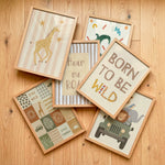 Six framed nursery art prints with light wood frames are arranged on a light wood floor. The prints feature safari-themed illustrations and playful text including 'BORN TO BE WILD', 'Hear me roar', 'STAY WILD', and 'STAY CHER'.