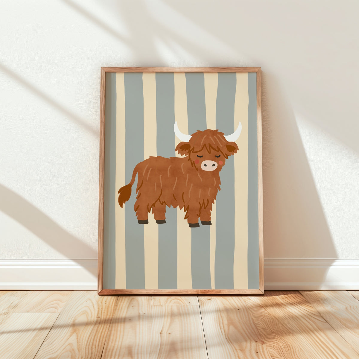 A framed art print featuring a cartoon Highland cow on a background of vertical beige and muted blue-gray stripes, leaning against a white wall on a light wood floor.