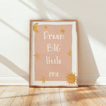 A framed nursery art print leaning against a white wall on a light wood floor. The print has a dusty pink background with the phrase 'Dream BIG little one' in white script, decorated with gold stars, a crescent moon, and a sun, in a light wood frame.