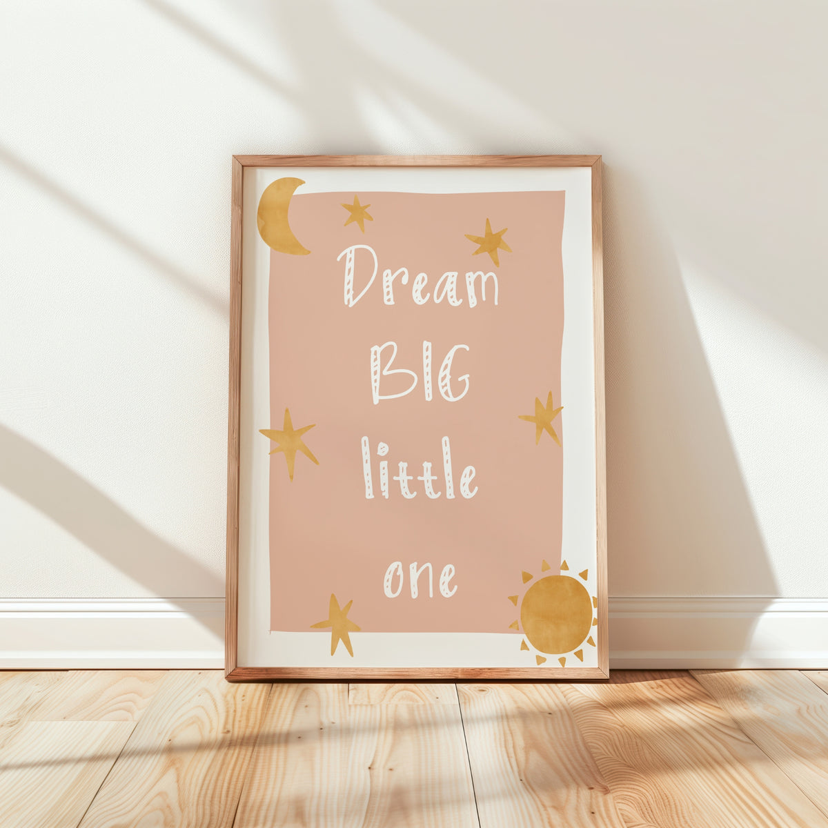 A framed nursery art print leaning against a white wall on a light wood floor. The print has a dusty pink background with the phrase 'Dream BIG little one' in white script, decorated with gold stars, a crescent moon, and a sun, in a light wood frame.