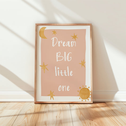 A framed nursery art print leaning against a white wall on a light wood floor. The print has a dusty pink background with the phrase 'Dream BIG little one' in white script, decorated with gold stars, a crescent moon, and a sun, in a light wood frame.