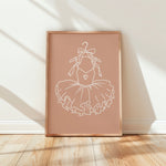 A framed minimalist art print featuring a white line drawing of a ballerina's tutu with a small heart on the chest, set against a dusty rose background. The print is housed in a light natural wood frame and is leaning against a white wall on a light wood floor, with soft diagonal shadows cast across the scene.