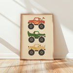 A framed vertical art print featuring three cartoon monster trucks in red, green, and tan on a cream background, leaning against a white wall on a light wood floor.