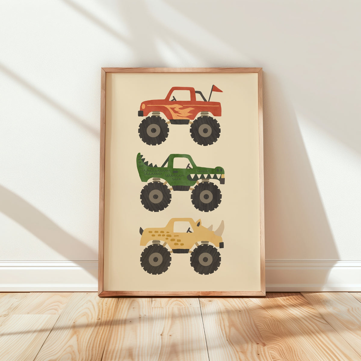 A framed vertical art print featuring three cartoon monster trucks in red, green, and tan on a cream background, leaning against a white wall on a light wood floor.