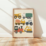 A framed children's art print featuring colorful construction vehicles, leaning against a white wall on a light wood floor with soft window shadows.