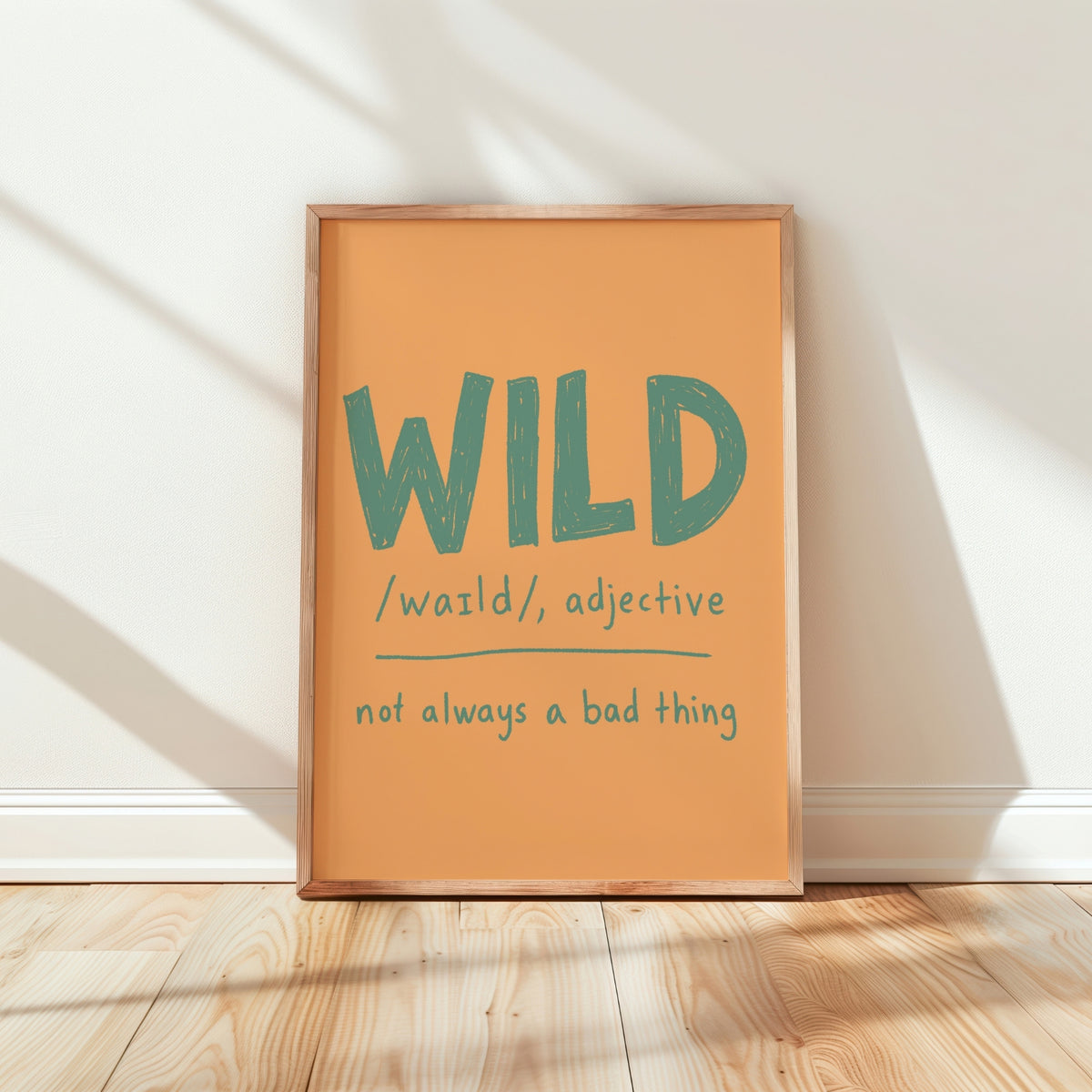 A framed typographic wall art print leaning against a white wall on a light wood floor. The print has a warm peach-orange background with the word “WILD” in large teal letters and smaller teal text reading “/waɪld/, adjective” and “not always a bad thing.