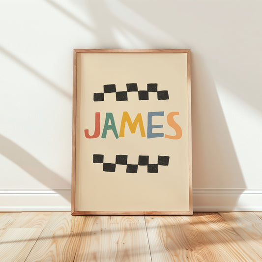 A framed personalized art print leaning against a white wall on a light wood floor. The print features the name 'JAMES' in colorful retro letters with black checkered bars above and below, set in a light wood frame.