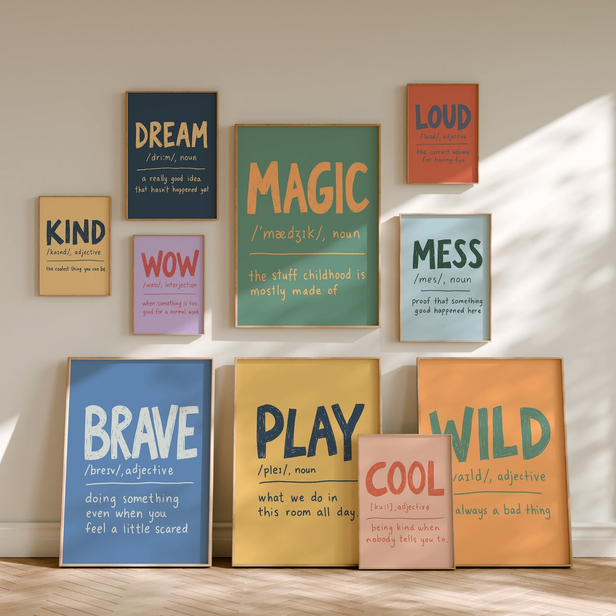 A set of framed typographic wall art prints arranged on a beige wall above a light wood floor. The prints feature colorful definitions of words such as DREAM, LOUD, KIND, WOW, MESS, BRAVE, PLAY, COOL, and WILD in a clean, modern aesthetic with warm natural light casting soft shadows.