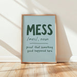 A framed typographic wall art print leaning against a white wall on a light wood floor. The print has a pale blue background with dark green text reading “MESS /mes/, noun proof that something good happened here” and is enclosed in a natural wood frame.