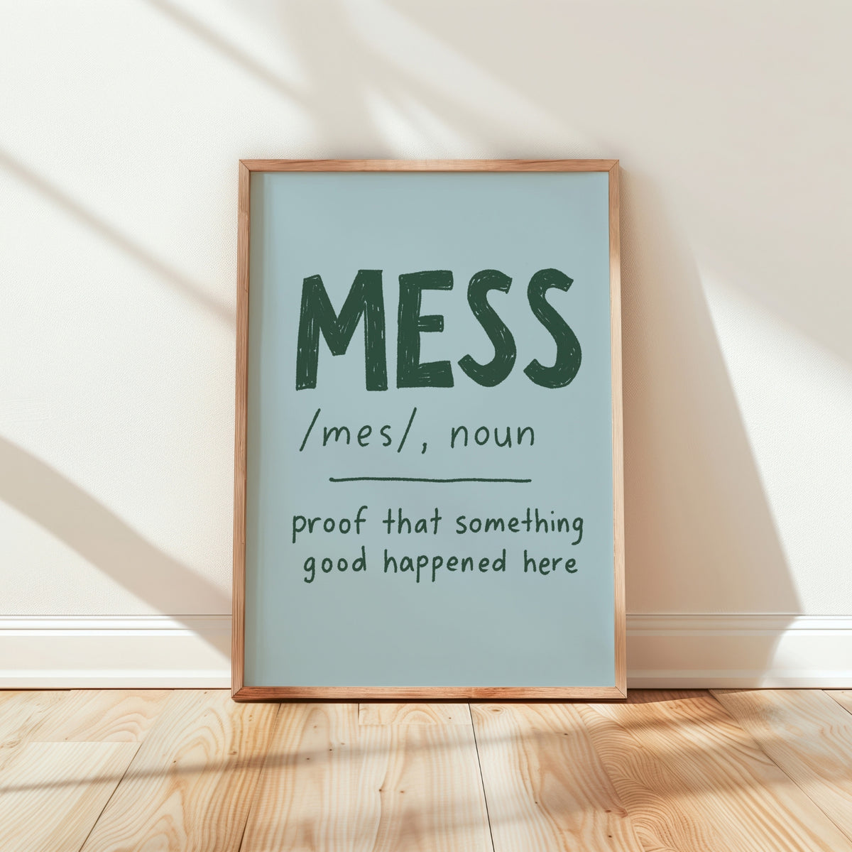 A framed typographic wall art print leaning against a white wall on a light wood floor. The print has a pale blue background with dark green text reading “MESS /mes/, noun proof that something good happened here” and is enclosed in a natural wood frame.