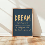 A framed typographic wall art print leaning against a white wall on a light wood floor. The print has a dark blue background with the word “DREAM” in large beige letters and a dictionary-style definition below.
