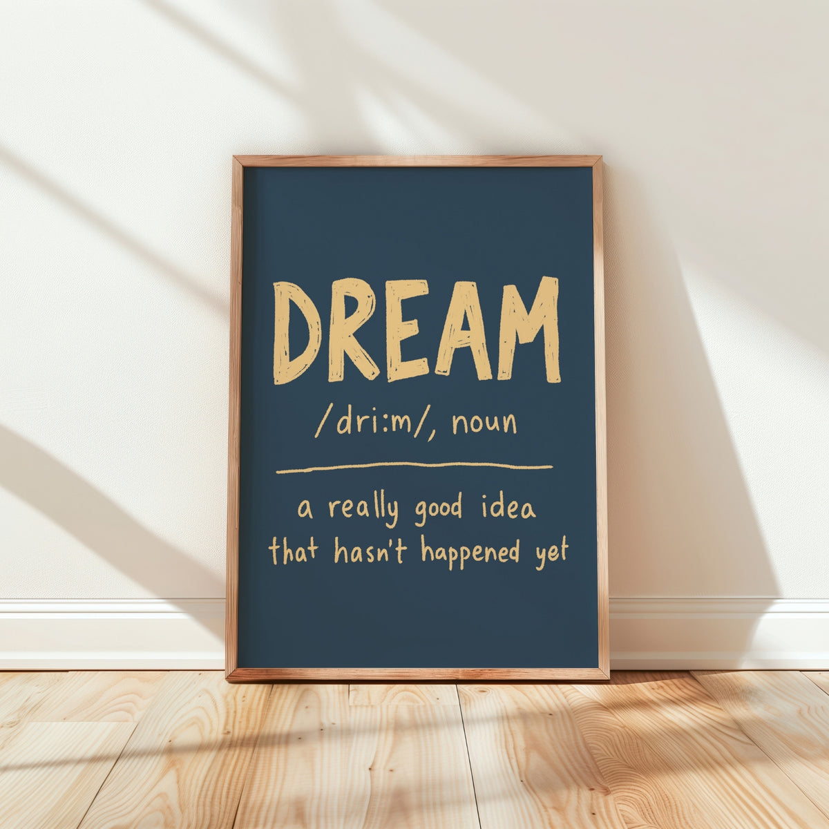 A framed typographic wall art print leaning against a white wall on a light wood floor. The print has a dark blue background with the word “DREAM” in large beige letters and a dictionary-style definition below.
