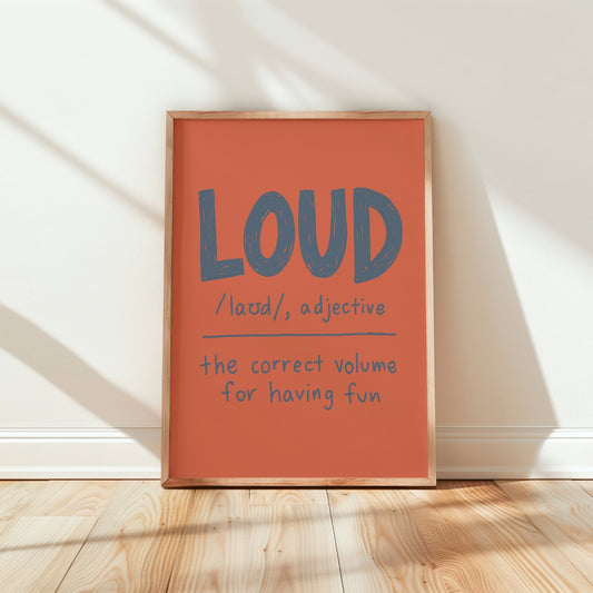 A framed typographic wall art print leaning against a white wall on a light wood floor. The print has a warm coral-orange background with large blue-gray text reading “LOUD” and smaller handwritten-style text below.