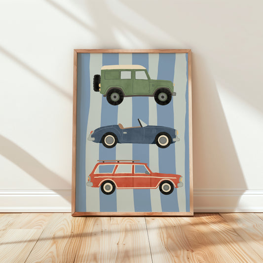 A framed art print leaning against a white wall on a light wood floor. The print features three retro cars stacked vertically on a background of light blue and white stripes: a green station wagon with a cream roof, a blue convertible, and a red-orange station wagon.