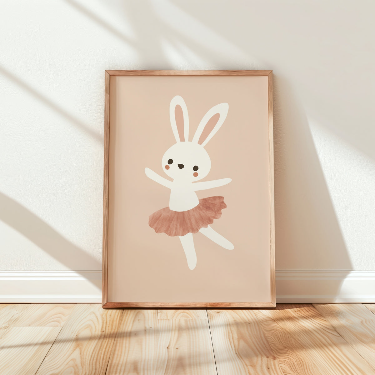 A framed children's art print featuring a white bunny ballerina with a dusty pink tutu, leaning against a white wall on a light wood floor with soft window shadows.