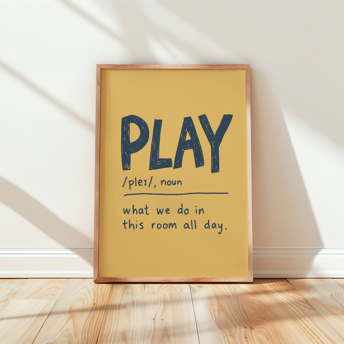 A framed typographic wall art print leaning against a white wall on a light wood floor. The print has a mustard yellow background with the word “PLAY” in large dark blue letters and a dictionary-style definition underneath.