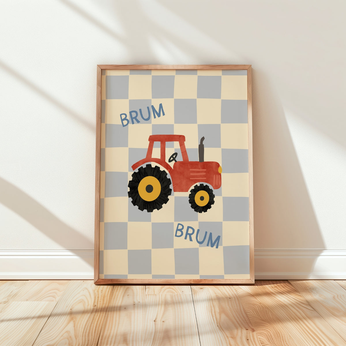 A framed children's wall art print featuring a red tractor on a cream and light gray checkerboard background with the word 'BRUM' repeated in blue. The framed print is leaning against a white wall on a light wood floor.